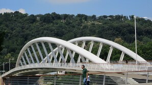 Un puente arqueado blanco con barandillas de metal, un hombre caminando sobre él y una exuberante ladera verde al fondo.