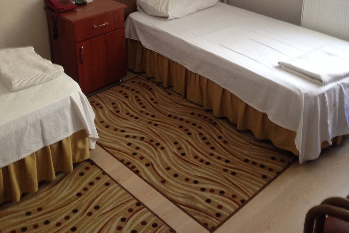 A small, single-bed room with a wooden headboard, a red bedside table, a patterned rug, and a wall-mounted light fixture.