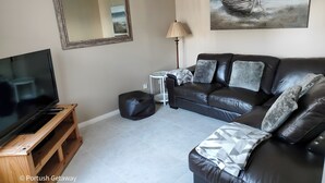 A living room with a TV on a wooden stand, a black leather sofa, a white side table, and a lamp. - Portrush Getaway – Family-Friendly Home Near Beaches & Town (Portrush)