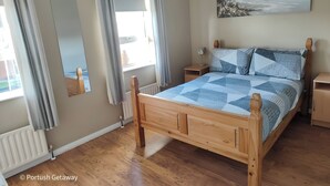 A wooden bed with a blue and white quilt, a bedside table, a nightstand, a mirror, and a painting of a seascape. - Portrush Getaway – Family-Friendly Home Near Beaches & Town (Portrush)