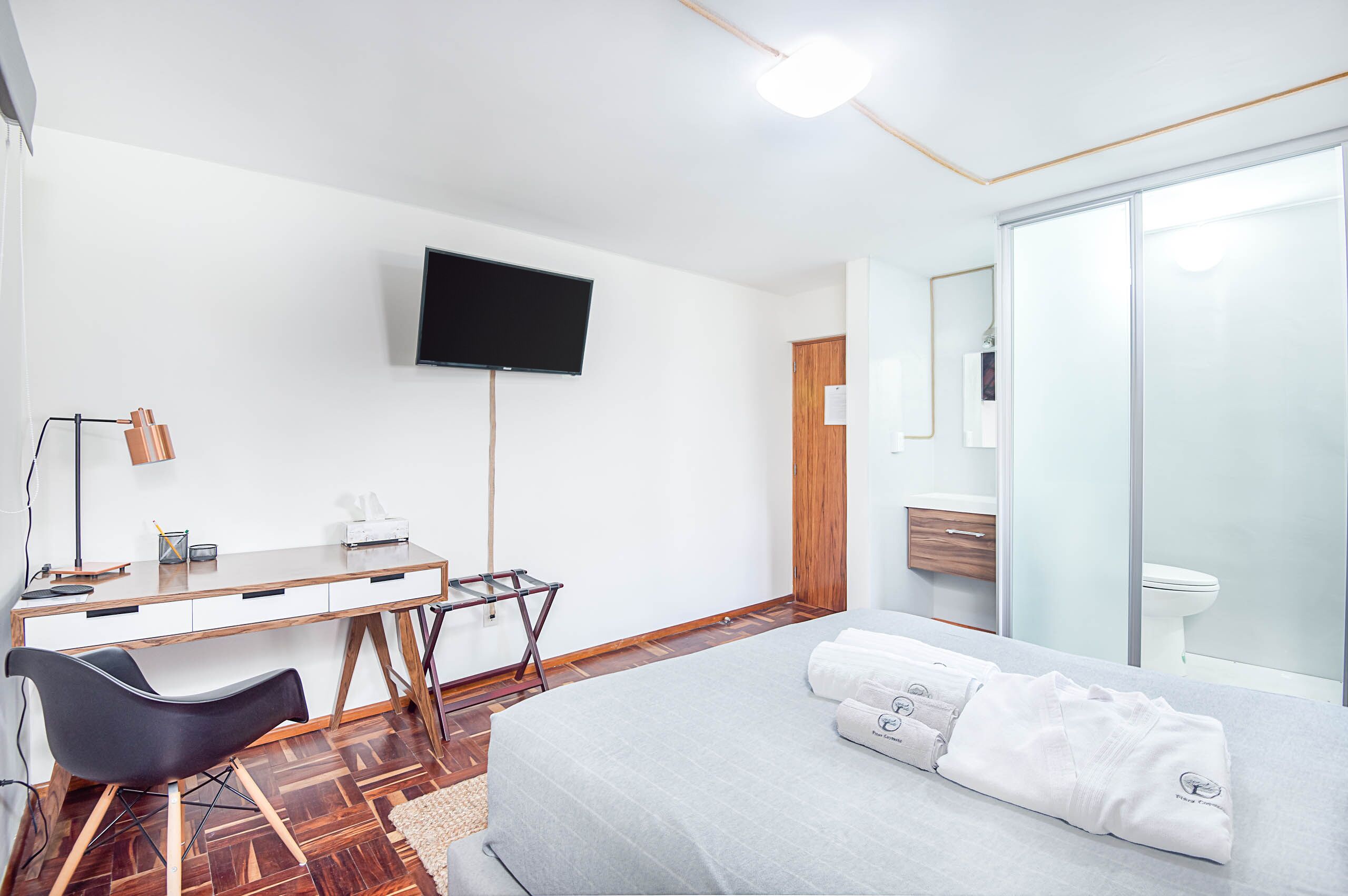 Habitación de hotel con cama, escritorio con silla, televisor de pantalla plana, piso de madera y baño con inodoro y lavamanos.