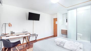 Habitación de hotel con cama, escritorio con silla, televisor de pantalla plana, piso de madera y baño con inodoro y lavamanos.