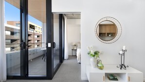 Une chambre d’hôtel moderne dotée d’une porte-fenêtre coulissante donnant sur un balcon, d’une console blanche avec un miroir et offrant une vue sur un immeuble situé de l’autre côté de la rue.