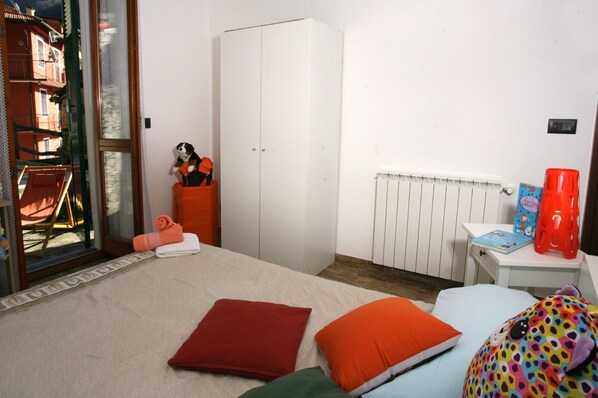 Un dormitorio con cama, armario, radiador, una mesita con un libro y un taburete rojo, y una ventana con vistas a edificios.