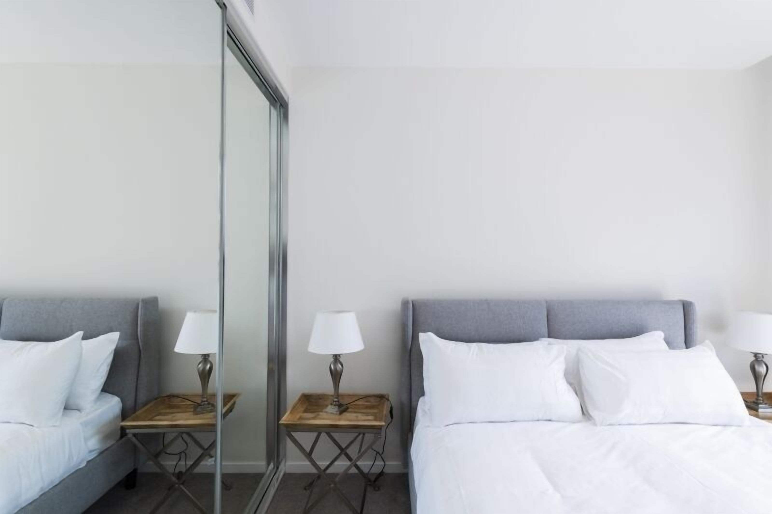 A hotel room with two beds, each with a grey headboard and white bedding. There are bedside tables with lamps and a large mirror on one wall.