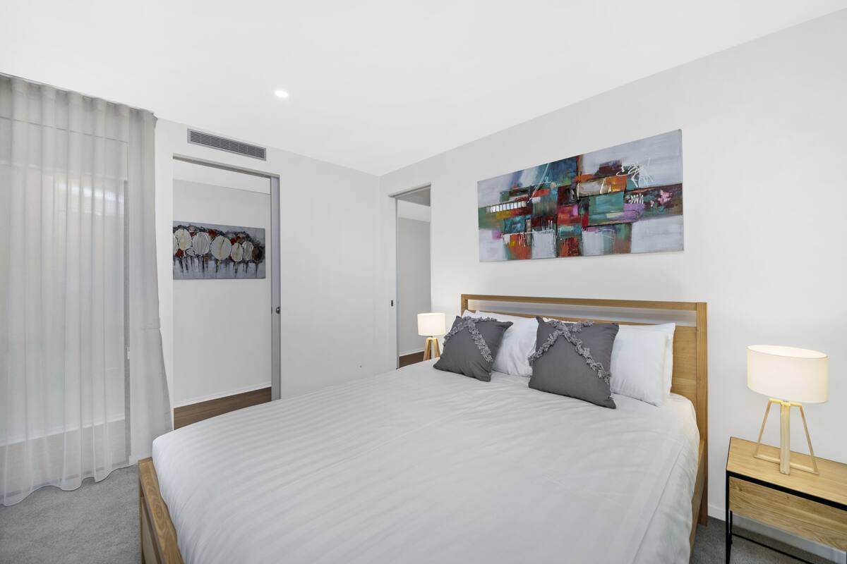 A modern hotel room with a large bed, two bedside tables with lamps, a wall-mounted abstract painting, and a door leading to another room.
