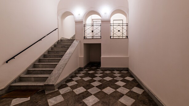 Un escalier avec un sol à carreaux, des murs blancs et des fenêtres en arc surmontées de grilles métalliques.