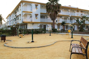 Ein Sandspielplatz mit Spielgeräten, einer Bank und im Hintergrund ein Gebäude mit Balkonen.