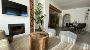Una sala de estar moderna con chimenea, televisor montado en la pared, comedor con mesa y sillas de madera, y una planta grande en maceta.