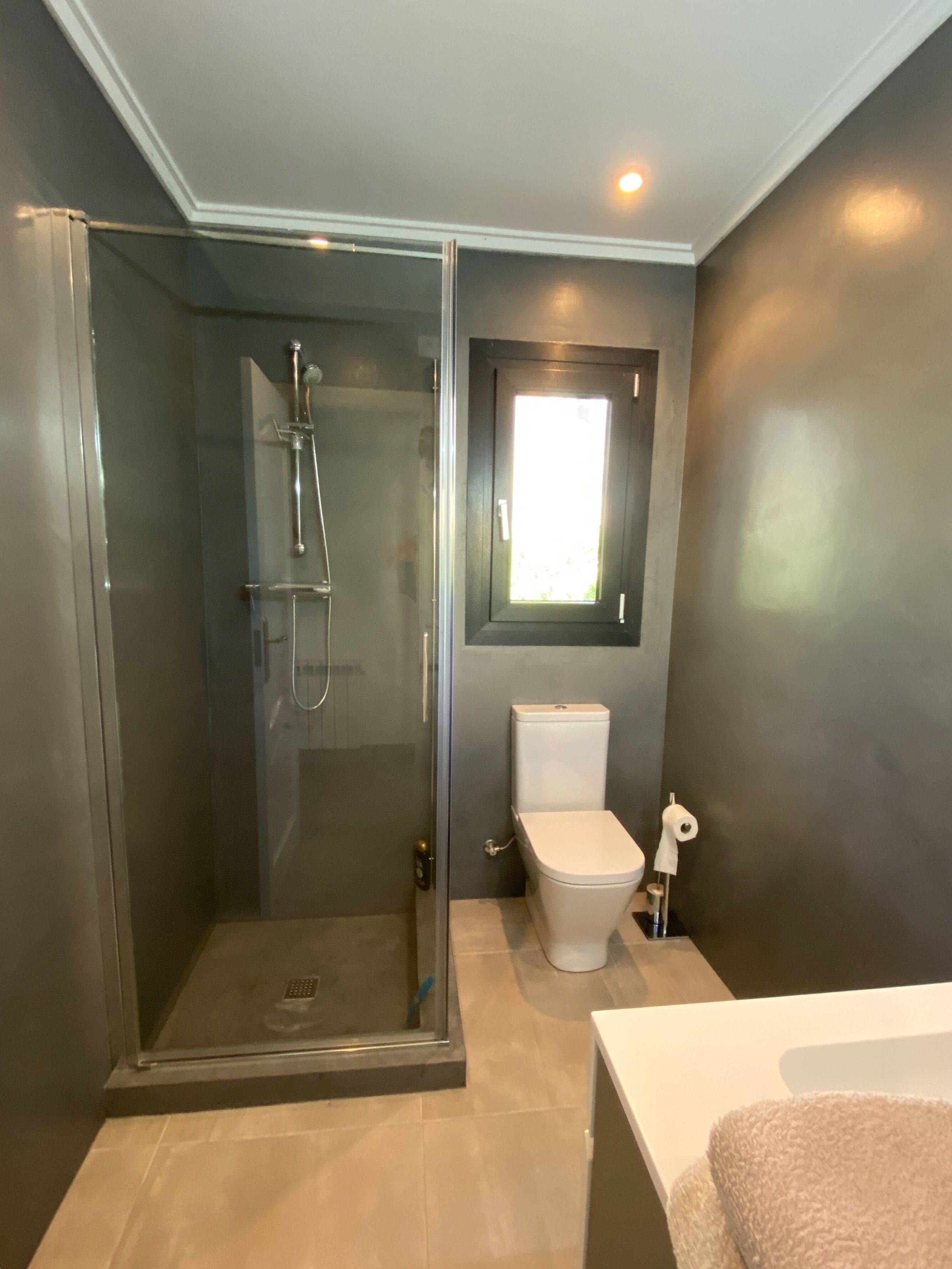 A modern bathroom with a glass-enclosed shower, a toilet, and a small window.