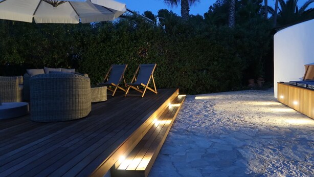 Terraza de madera con mobiliario exterior, que incluye un banco y sillas, bajo un gran paraguas.