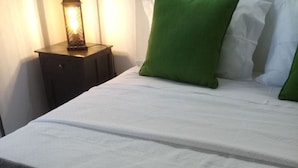 Una cama bien hecha con sábanas blancas y almohadas verdes, una mesita de noche oscura con una lámpara, y toallas grises en el suelo.
