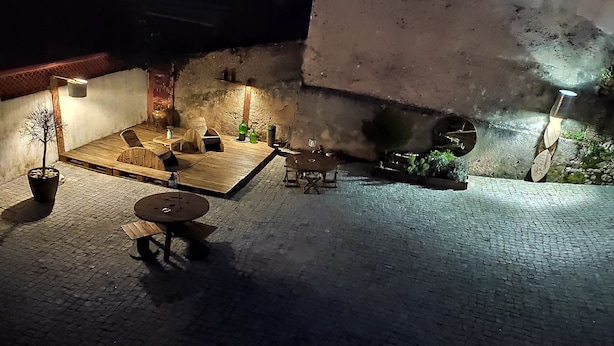 Un patio al aire libre con mobiliario de madera y plantas en macetas, iluminado con luces colgantes.