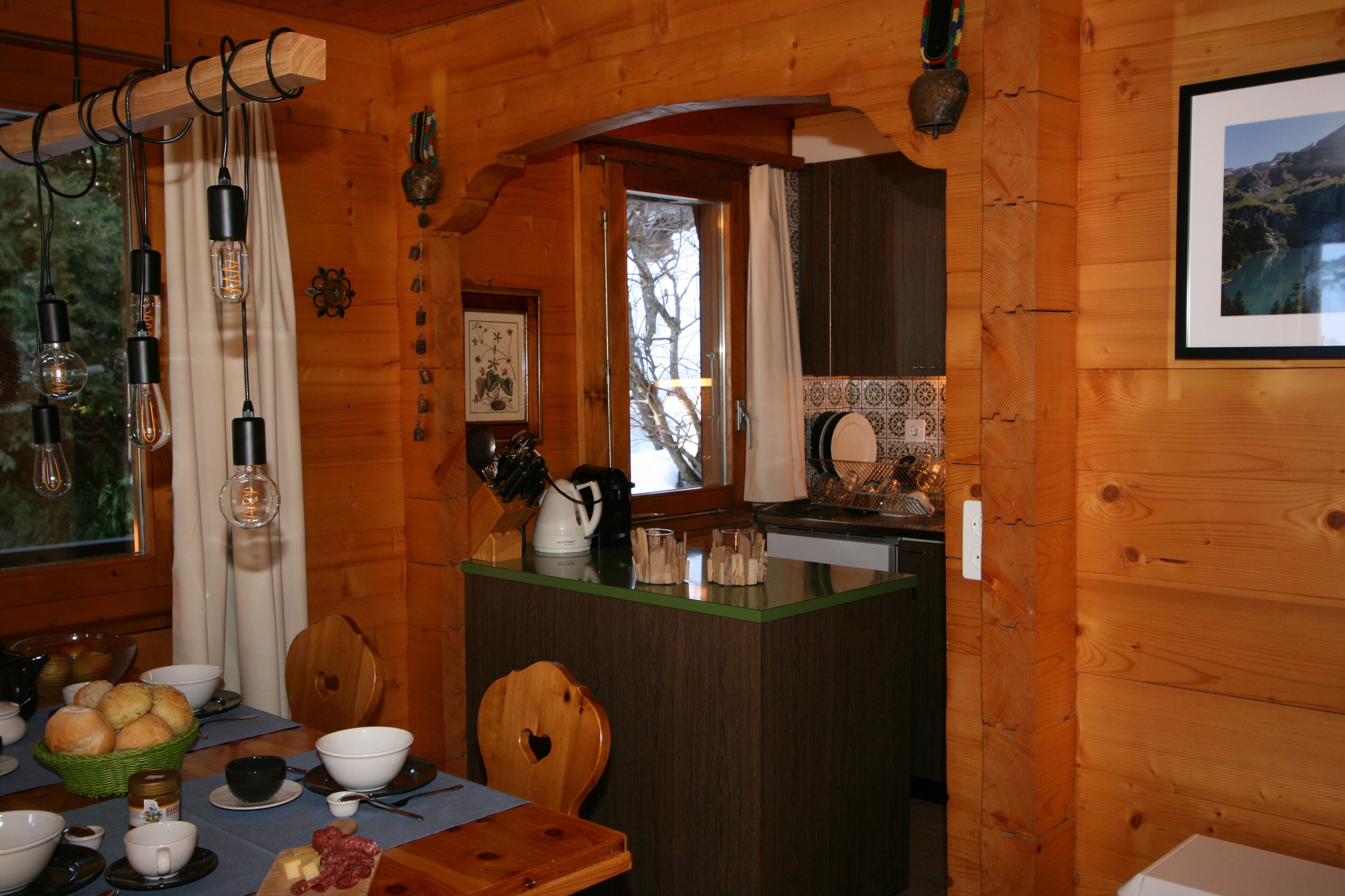 L’intérieur d’un chalet en bois présente une table à manger dressée pour le petit-déjeuner, un coin cuisine et une fenêtre donnant sur les arbres.