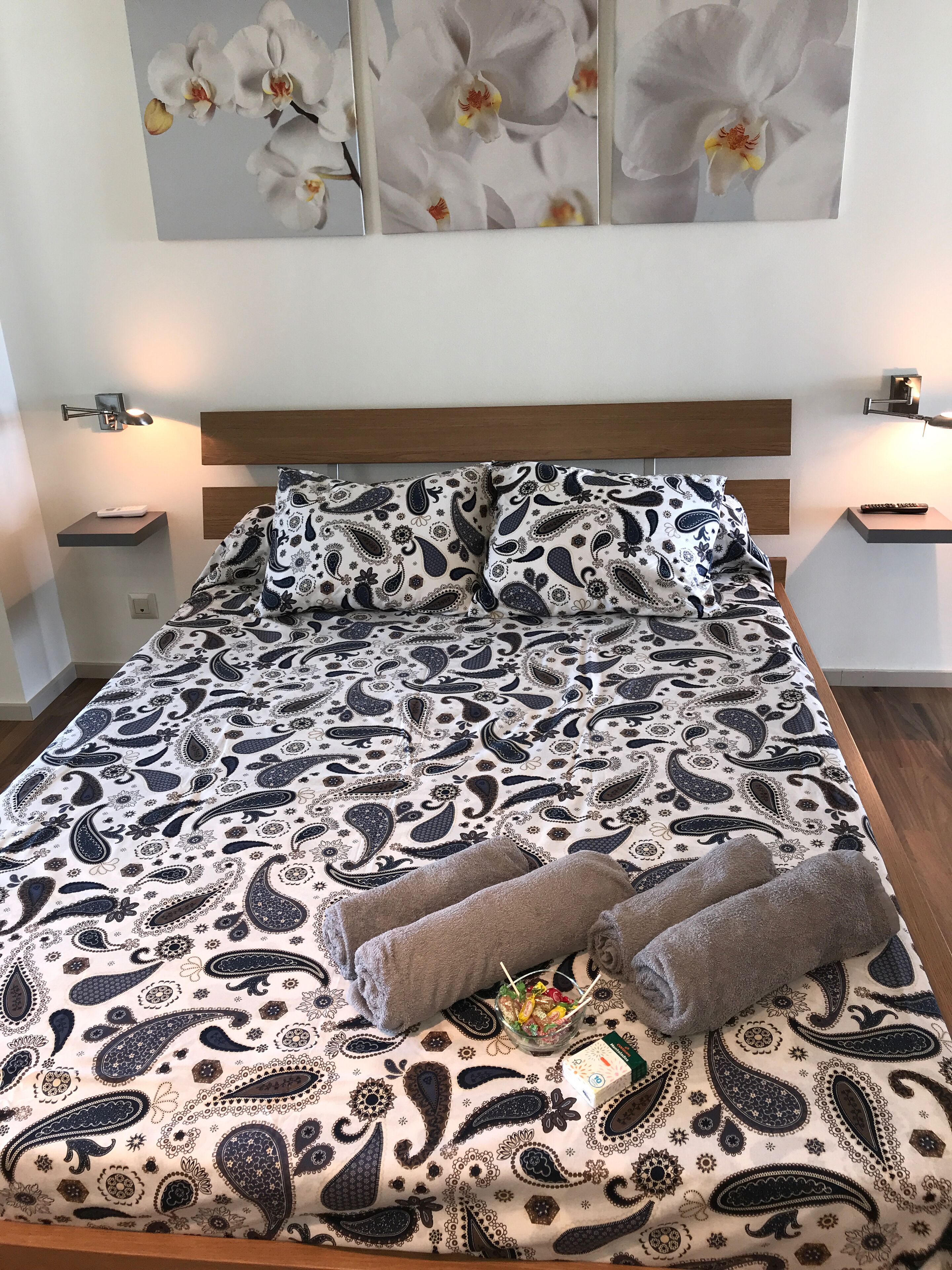 A bed with a patterned bedspread, two pillows, a bedside table with a lamp, and a wall with a floral painting.