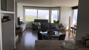 Un salon moderne avec un canapé gris, une table basse en verre et une vue sur la mer grâce à de grandes fenêtres.