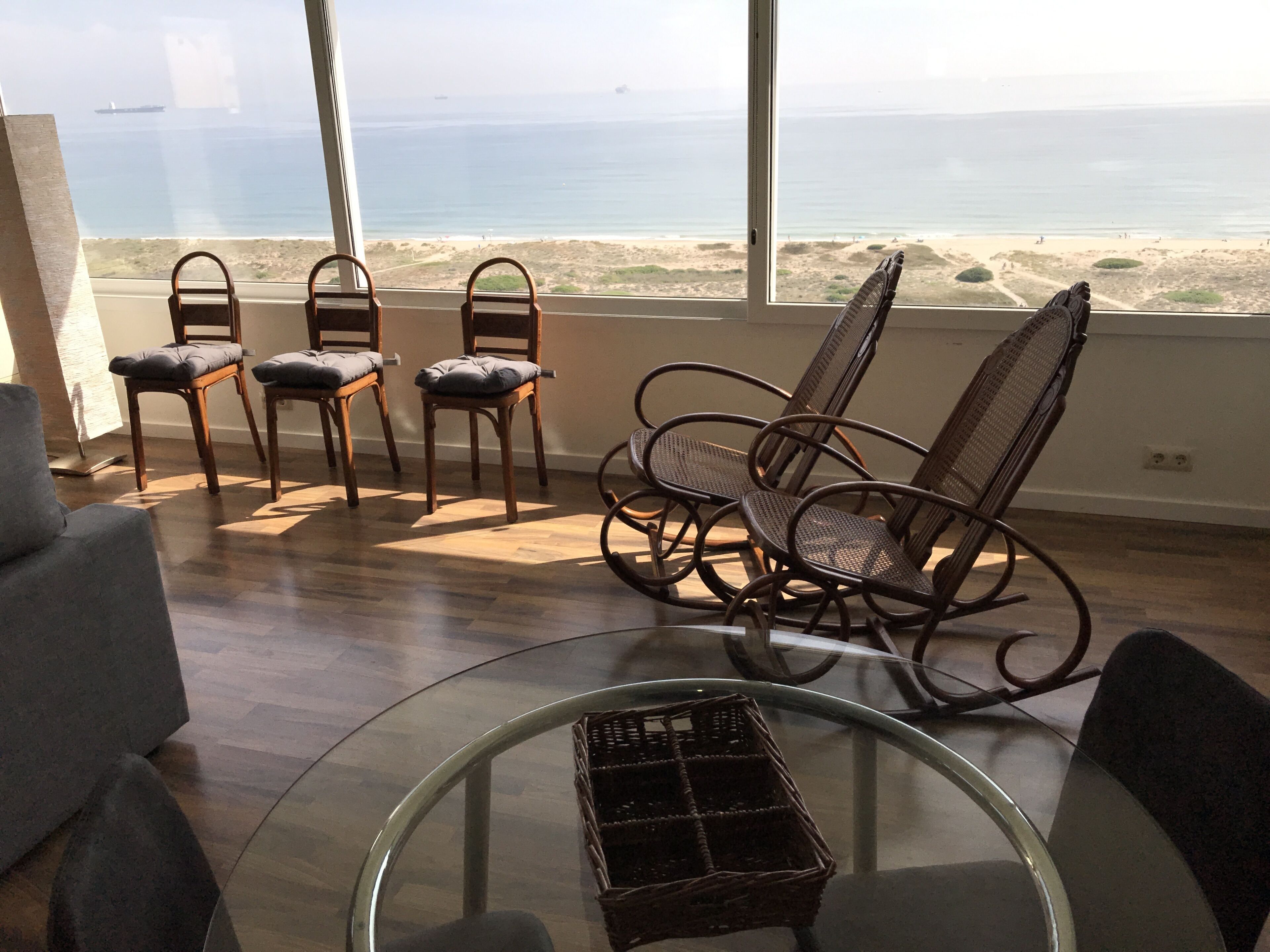 A room with a large window overlooking a beach, wooden chairs, a glass table, and a wicker basket.