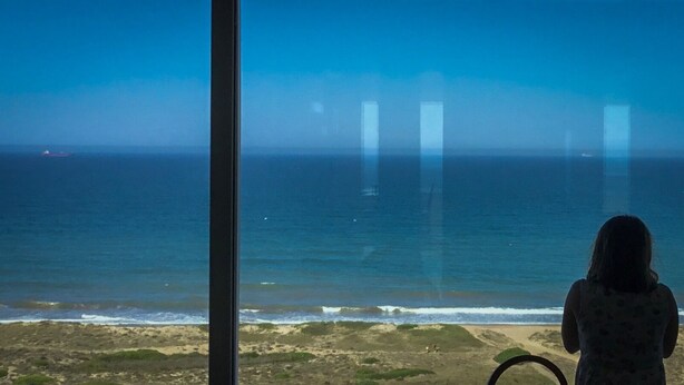 Une personne se tient près d’une fenêtre donnant sur une plage, avec vue sur l’océan et un navire à l’horizon.
