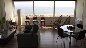 Un vaste espace de vie doté d’une grande fenêtre donnant sur une plage, et aménagé avec un canapé, des fauteuils et une table à manger.