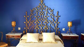 Une chambre avec un mur bleu, une tête de lit décorative en métal, deux tables de chevet avec des lampes, une chaise en bois et un lit avec une literie blanche.