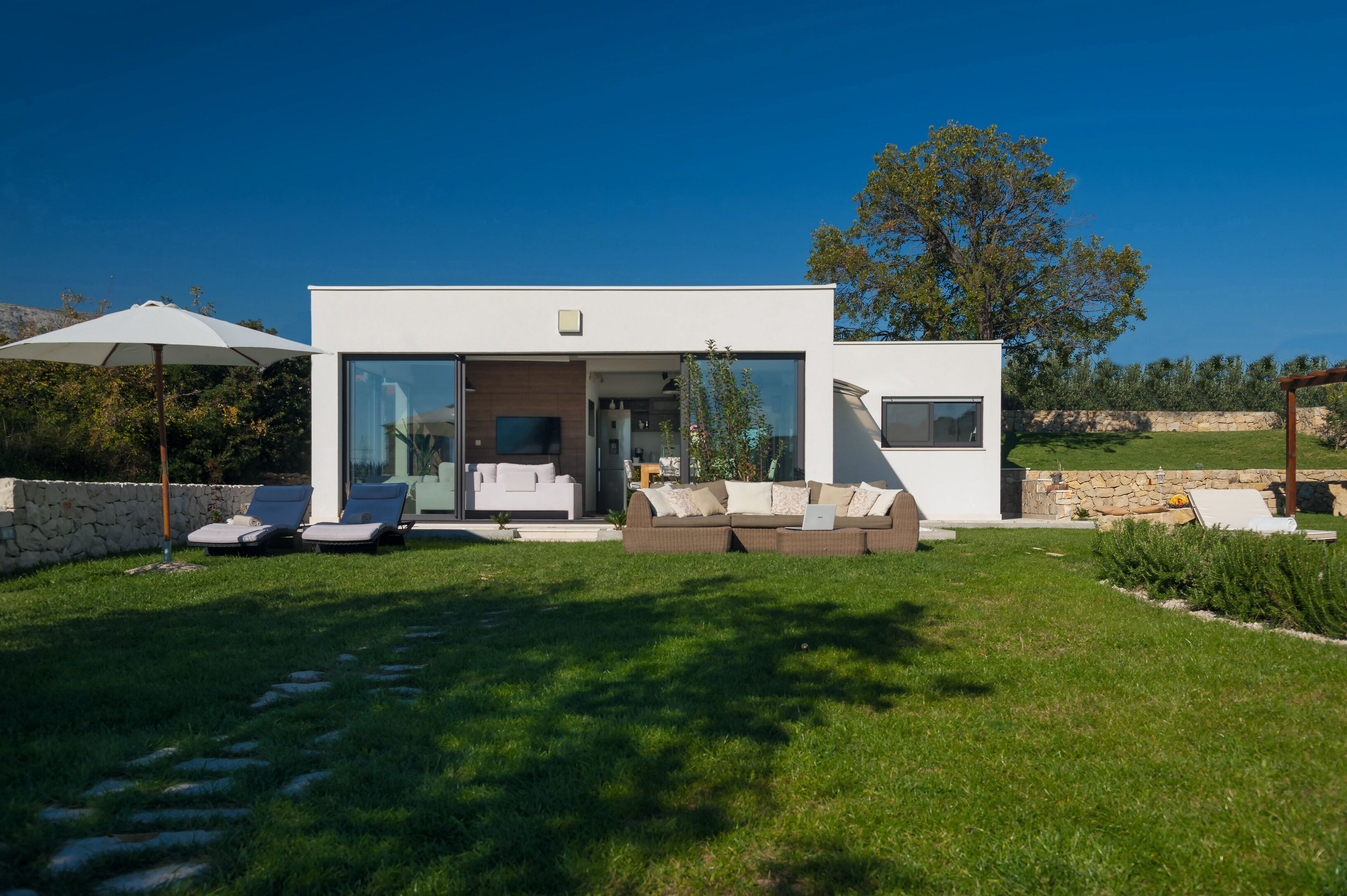 Une maison moderne avec une entrée dotée d’une porte en verre, un patio avec des fauteuils et un mur en pierre.