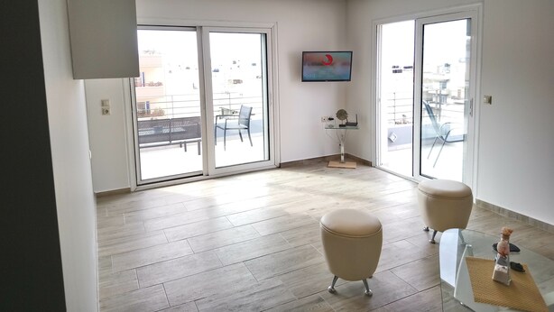 A modern living room with a large sliding glass door leading to a balcony, a flat-screen TV mounted on the wall, and a small table with decorative items. - Limena's penthouse apartment (Limenaw Hersonisou Crete)