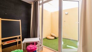 Una habitación con una pared negra, una mesa blanca, un taburete rosa y otro verde, además de una estantería de madera con libros.