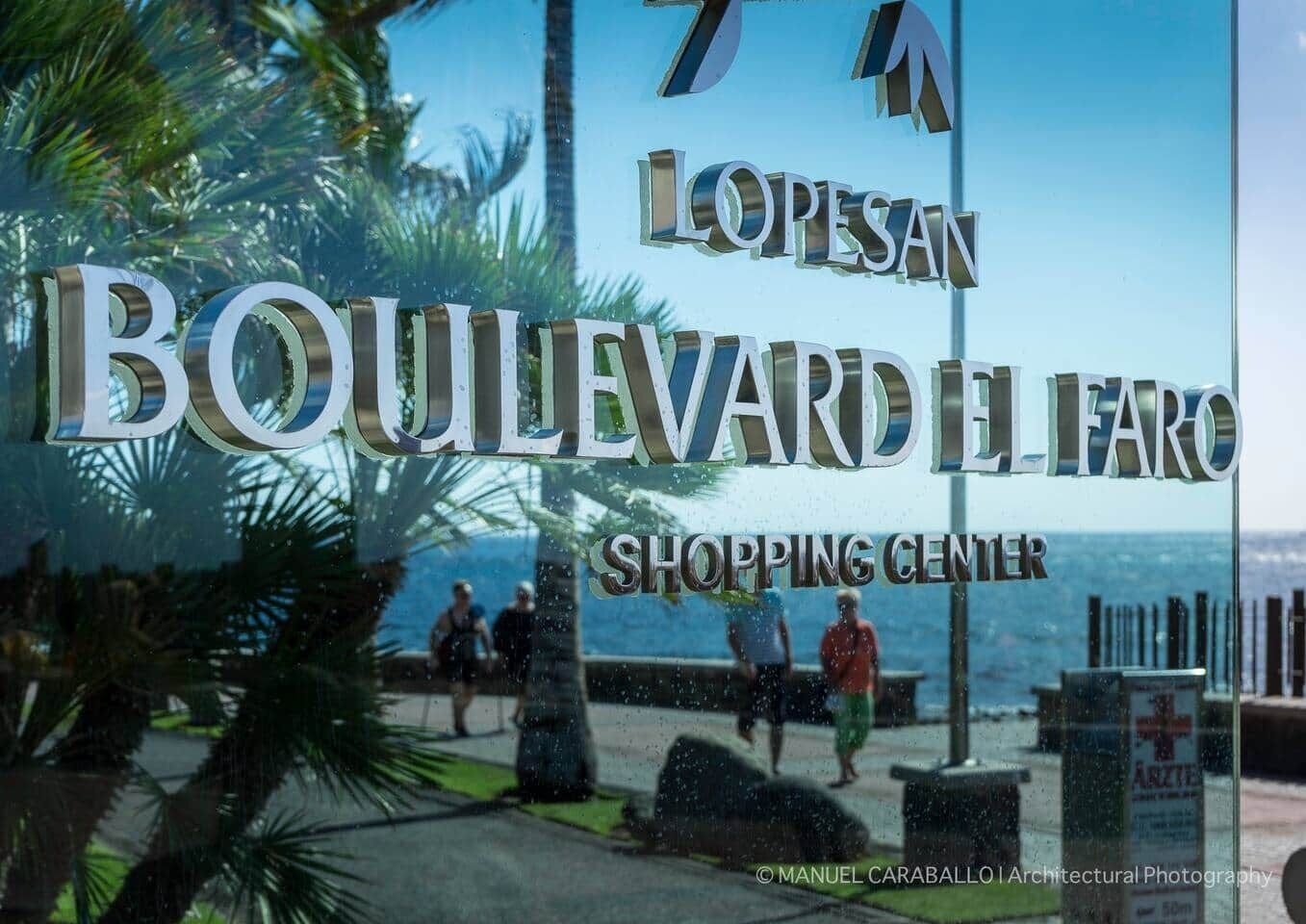A sign for 'Boulevard El Faro Shopping Center' with palm trees and a view of the ocean in the background.