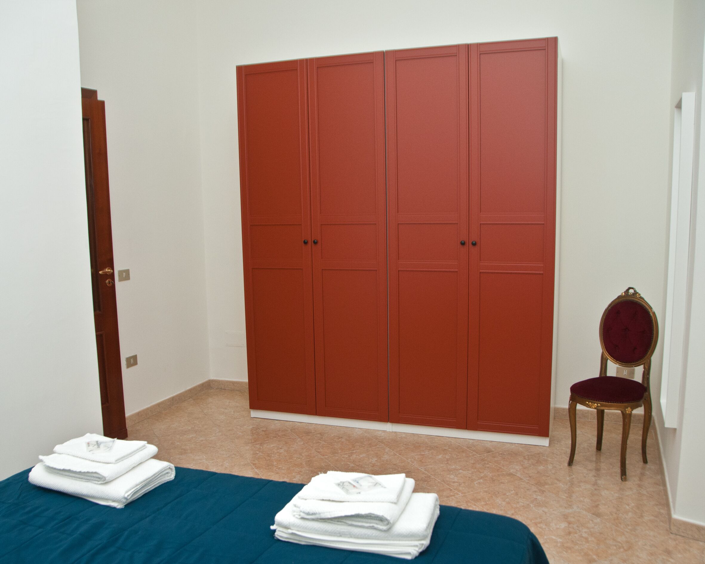 A room with a large red wardrobe, a wooden chair, and a bed with folded white towels.