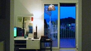 Ein Zimmer mit weißem Schreibtisch, einem Stuhl, einer Lampe und Blick durch eine Schiebetür nach draußen.