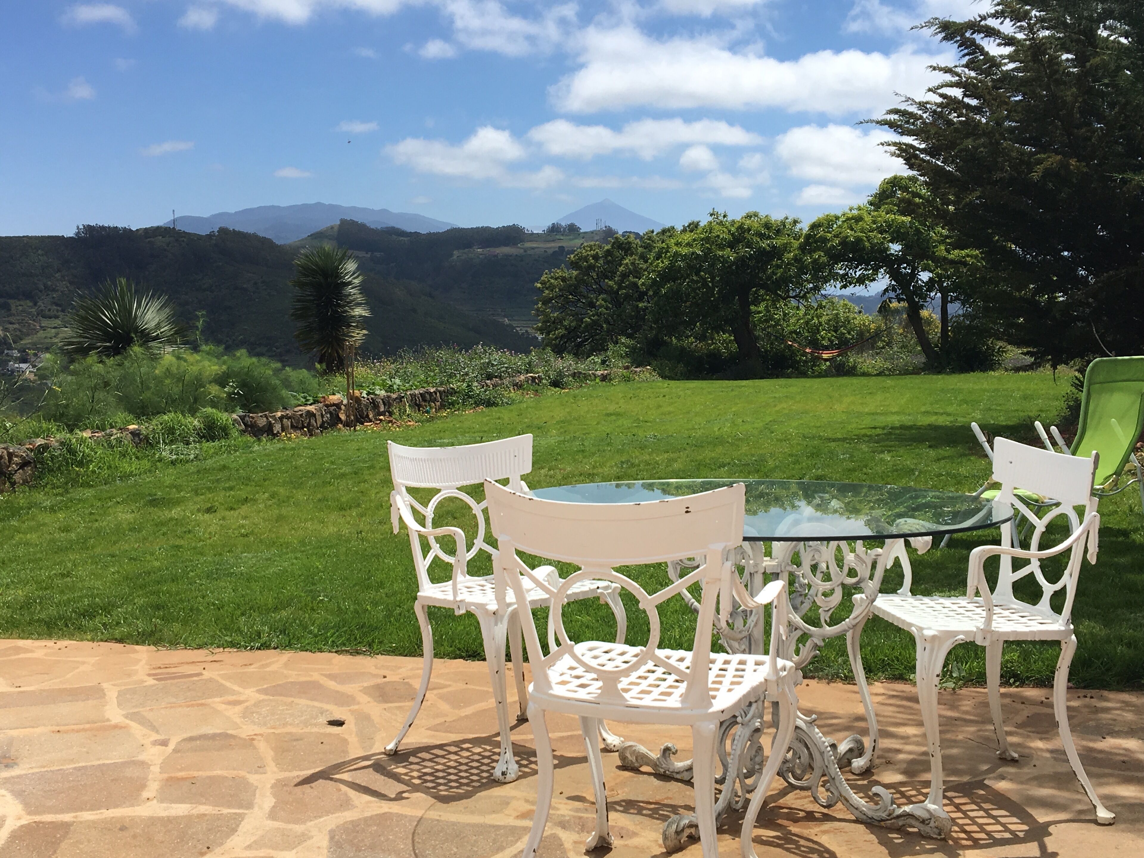 Un patio con una mesa de vidrio y sillas de metal blanco, con vista a un paisaje cubierto de césped, árboles y montañas a lo lejos.