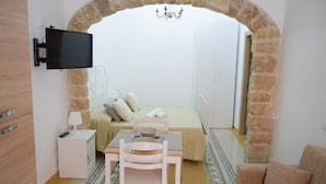 A bedroom with a stone archway, a bed, a desk with a chair, a TV mounted on the wall, and a chandelier.