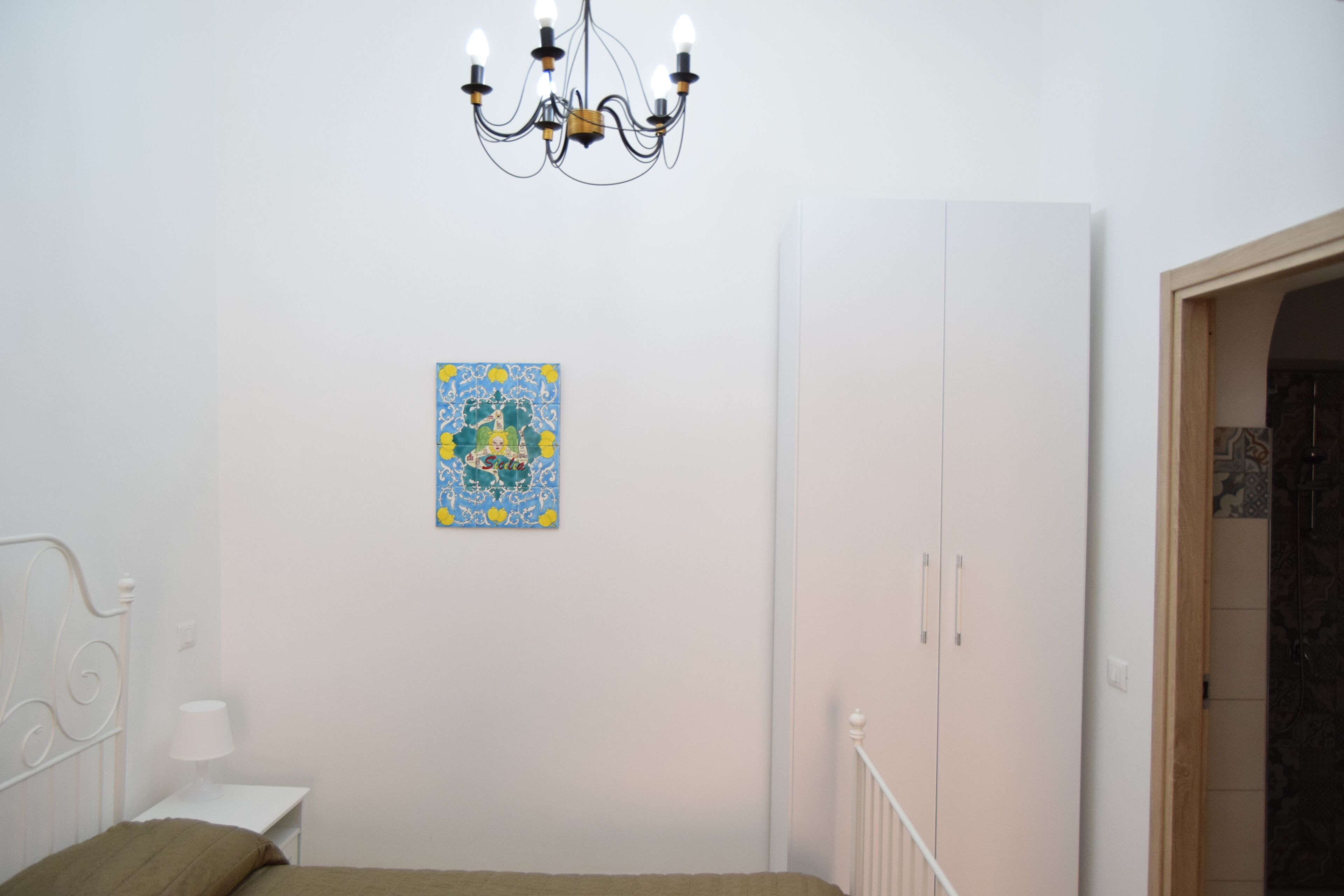 A bedroom with a bed, a white nightstand, a chandelier, a wardrobe, and a colorful painting on the wall.