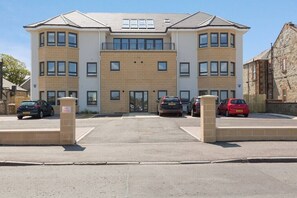 A multi-story residential building with a central entrance, parking spaces, and a stone wall on the right.