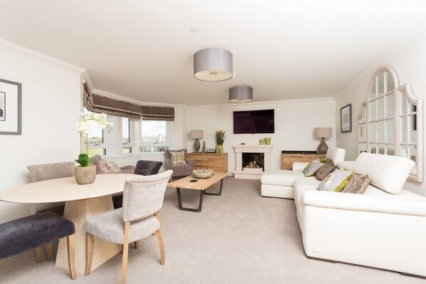 A modern living room with a dining area, white sofas, a fireplace, and a large window.