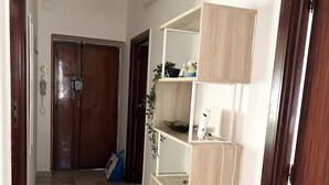 A hallway with a stone-tiled floor, a wooden door, a white pendant light, and a built-in shelving unit with various items. - Cozy and spacious apartment (Alghero)