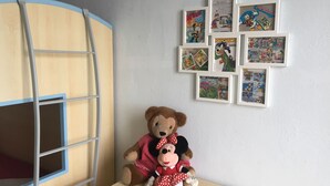Quarto com uma cama de solteiro, uma pequena mesa com um ursinho de pelúcia e um boneco do Mickey e da Minnie, e uma parede decorada com páginas de histórias em quadrinhos emolduradas.