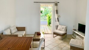 Una habitación con una mesa de madera, un sofá, una silla, un televisor y vista a un jardín por una puerta.