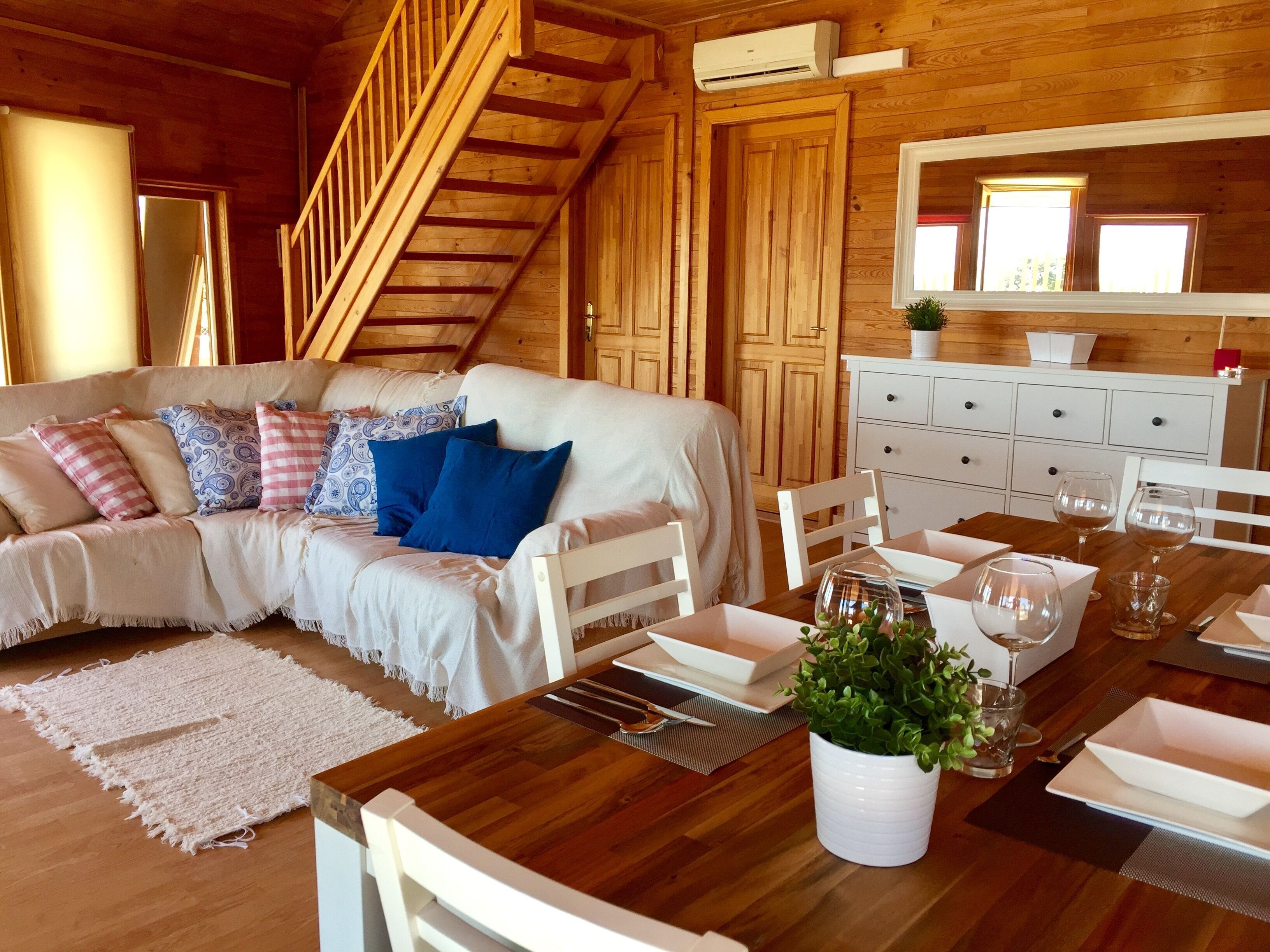 A cozy living room with a wooden staircase, a white sofa with patterned cushions, a dining table set with white plates and glasses, and a potted plant.