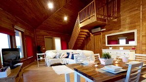 Ein gemütliches Blockhaus-Interieur mit einer Holztreppe, einem Bett mit Kissen, einem Esstisch gedeckt mit Gläsern und einer Pflanze sowie einem Fernseher auf einem Lowboard.