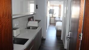 Une cuisine compacte équipée d’un grille-pain, d’un évier et de placards. Un couloir mène à une chambre à coucher comprenant un lit et une table de chevet.