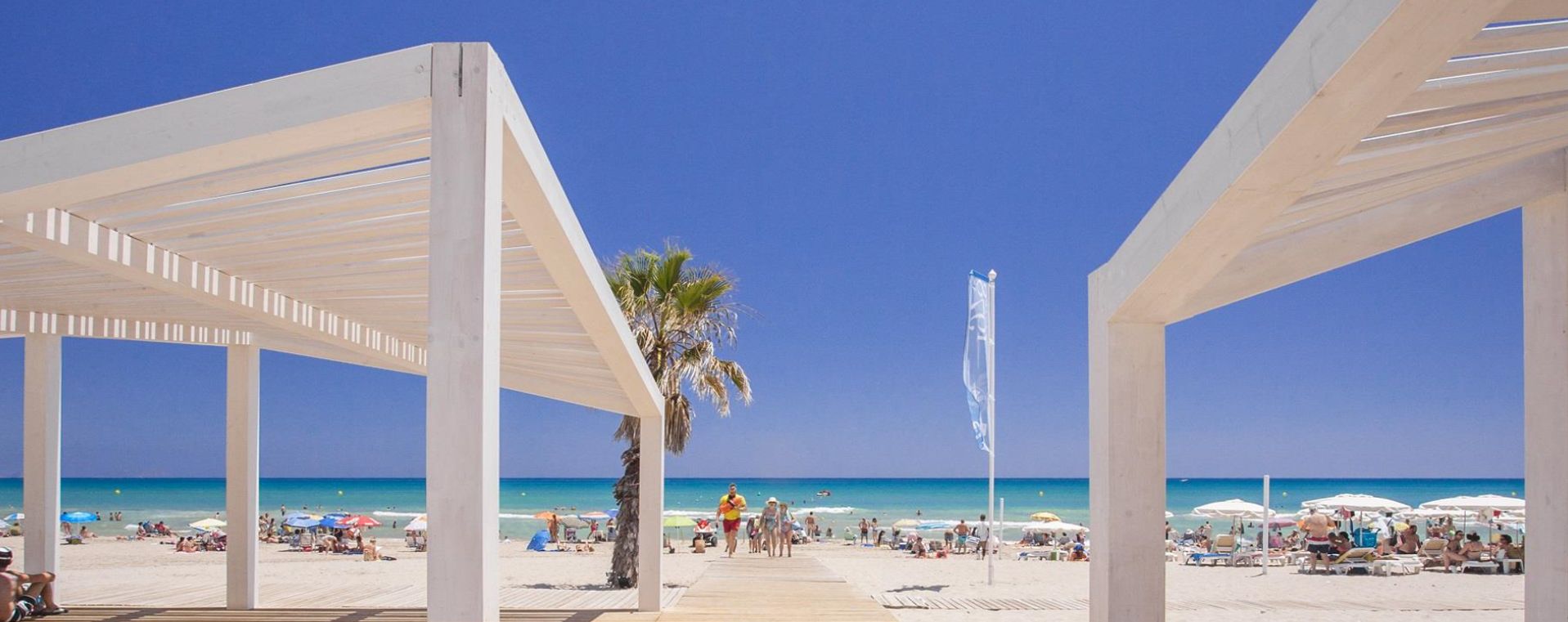 Una playa con un paseo de madera que lleva a la orilla, palmeras y sombrillas.