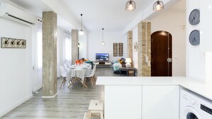 Cozinha moderna com máquina de lavar embutida, espaço de jantar com mesa comprida e sala de estar com televisão e assentos.