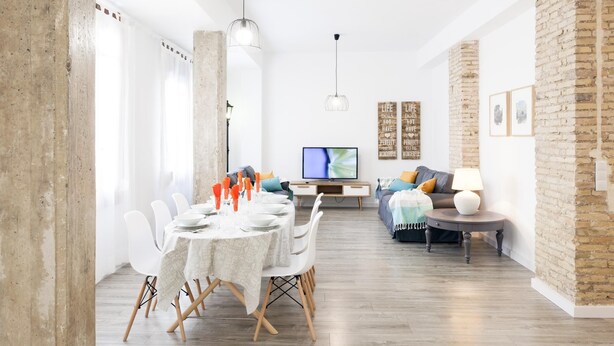 Sala de estar moderna com área de jantar, apresentando uma parede de tijolos, um sofá, uma TV e uma mesa posta para seis pessoas.