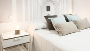 Quarto com cama branca, várias almofadas, mesa de cabeceira com abajur e parede branca com janela.
