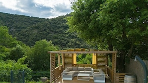 Una terraza de madera con un sofá integrado y una mesa de centro, rodeada por una pérgola de madera, con un fondo de exuberante vegetación y colinas.