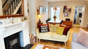 Een woonkamer met een open haard, een bank met opvallende kussens, een eethoek met een tafel en stoelen, en een keuken op de achtergrond.