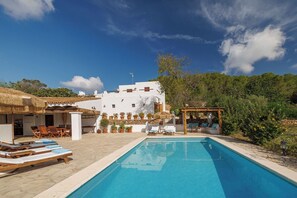 A pool area with lounge chairs, a pergola, and a white building with a thatched roof.