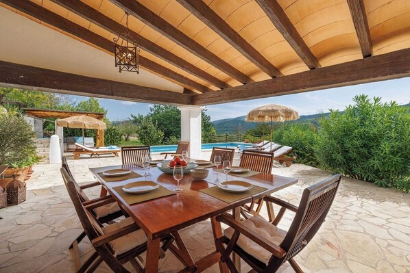 A covered outdoor dining area with a pool, wooden furniture, and a view of the mountains.