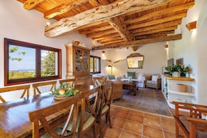 A rustic interior with a wooden dining table, chairs, a sofa, a cabinet, and a view through a large window.